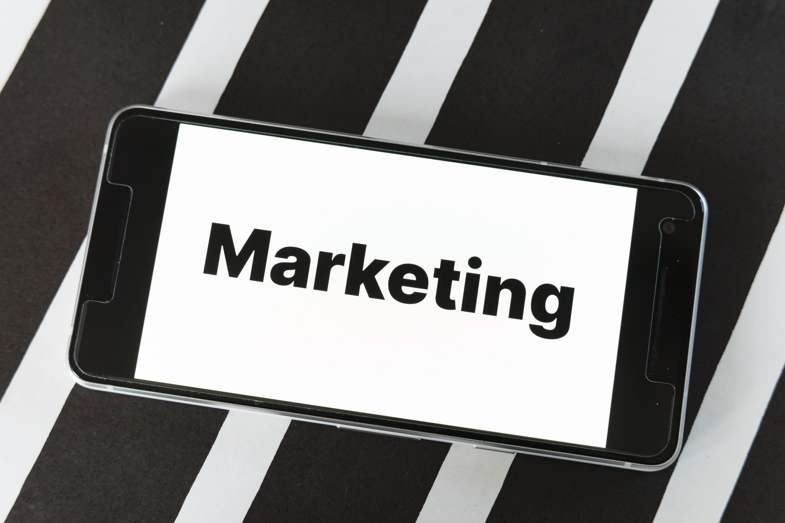 A smartphone displays the word “Marketing” in bold black text on a white screen, placed against a striking background of alternating black and white diagonal stripes. The image visually emphasizes digital marketing, communication, and branding themes, making it suitable for business presentations, advertising content, or mobile-focused promotional materials.