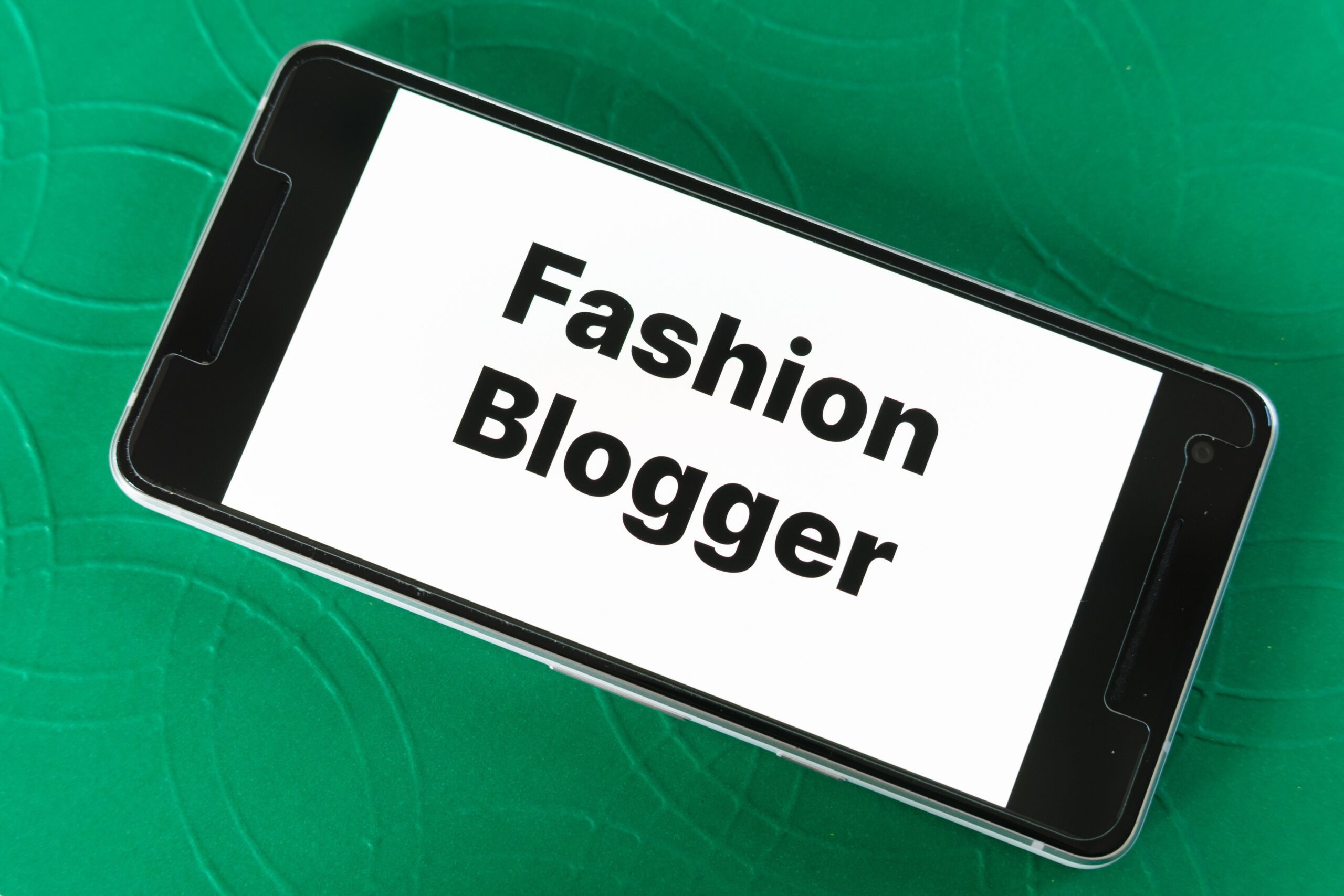 Smartphone displaying "Fashion Blogger" on screen, placed on a green textured surface.
